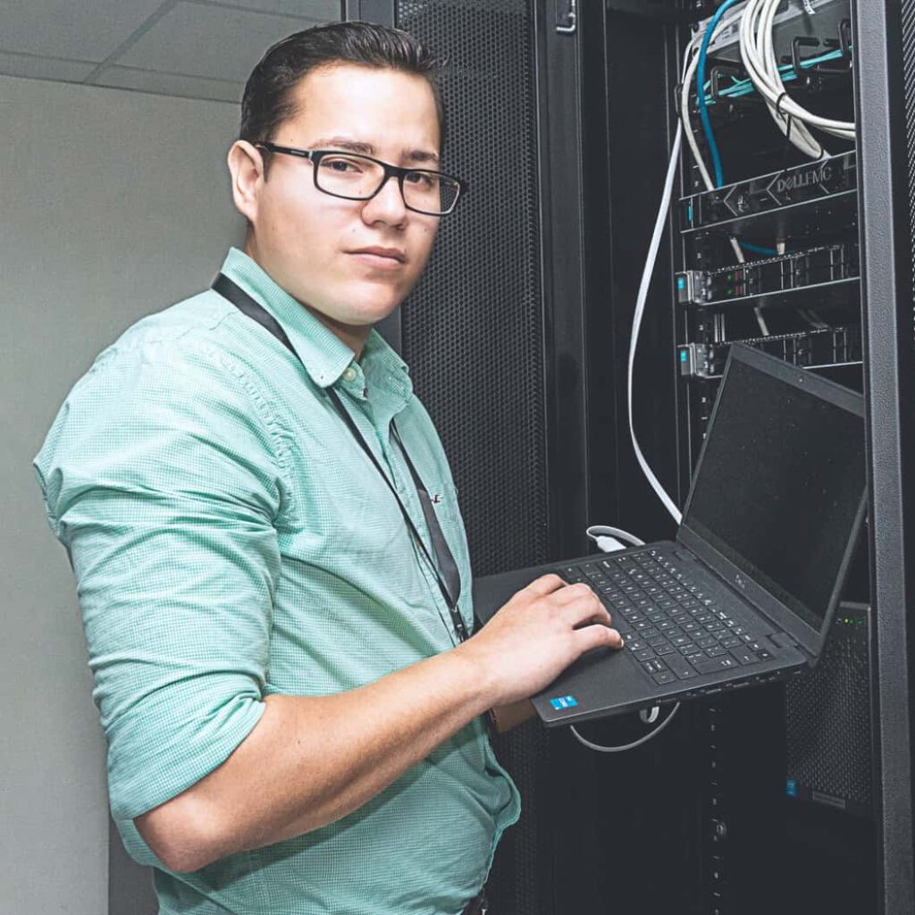 EDUCACIÓN CONTINUA Persona con gafas y camisa verde clara trabajando con un computador portátil frente a un rack de servidores. La escena muestra equipos de red y cableado organizado dentro del gabinete, mientras la persona opera el teclado del portátil en un entorno tecnológico o de centro de datos.